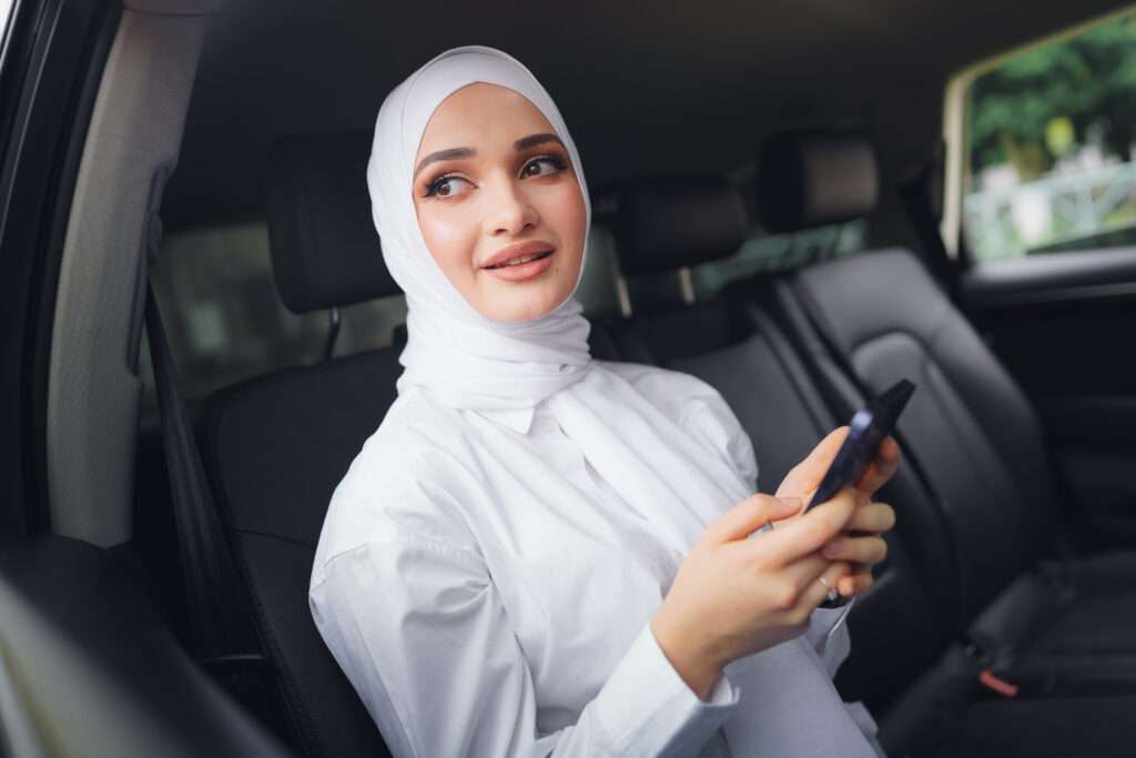 Beautiful muslim woman wearing white hijab sitting on the back seat of a car and using smartphone