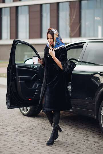 full length fashionable lady black coat leather boots wearing colorful handkerchief around her neck she is carrying cup takeaway coffee against open car street